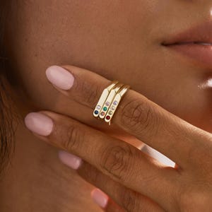 Puede incluir: Tres anillos dorados con barras rectangulares planas, cada uno con una pequeña piedra preciosa redonda. Las piedras son azules, verdes, rojas, blancas y moradas, dispuestas en una pila vertical en un dedo con esmalte de uñas rosa claro.