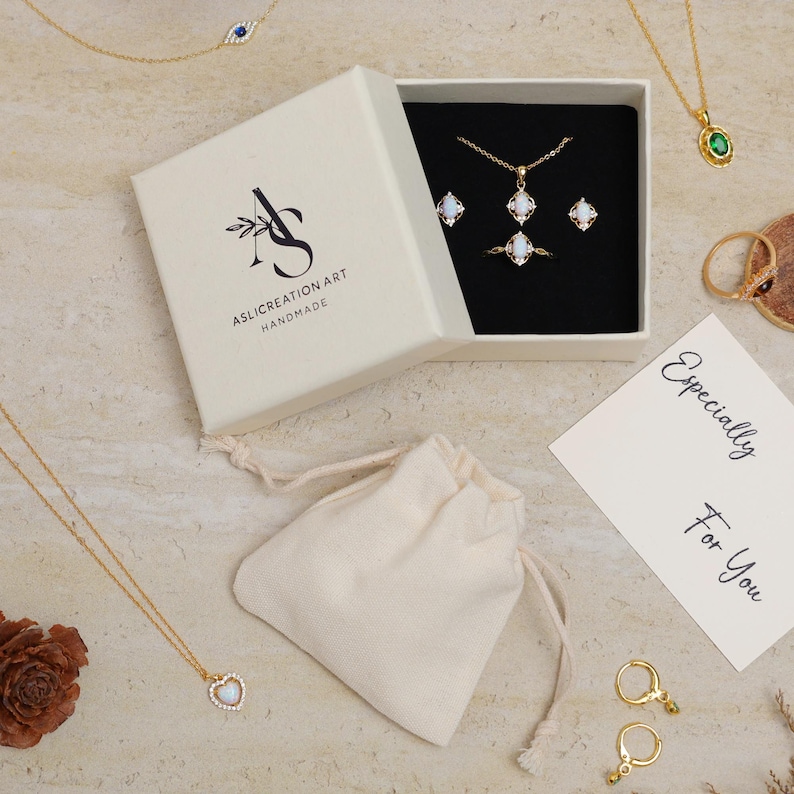 May include: A jewelry set in a white box with a black interior, featuring a necklace, ring, and earrings with opalescent stones. A small drawstring bag and a card that says "Especially For You" are also present. Other gold necklaces and rings are visible.
