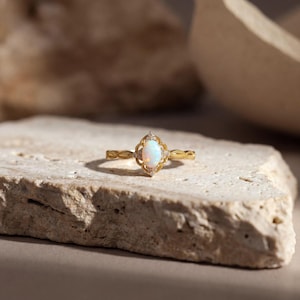 May include: A gold ring with an oval, iridescent opal center stone. The ring features a filigree design around the opal and small accent stones. The band has a delicate, textured pattern. The ring is displayed on a light-colored, textured stone surface.