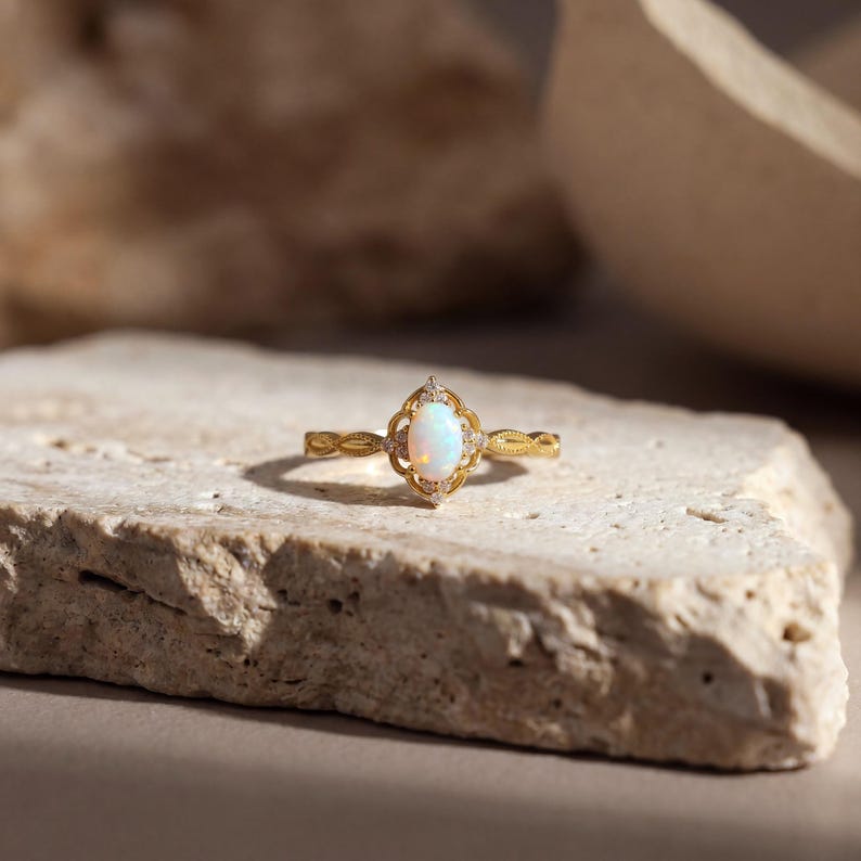 May include: A gold ring with an oval, iridescent opal center stone. The ring features a filigree design around the opal and small accent stones. The band has a delicate, textured pattern. The ring is displayed on a light-colored, textured stone surface.