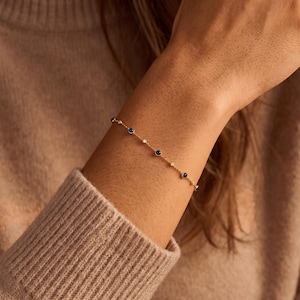 Puede incluir: Una delicada pulsera de cadena dorada con una serie de peque&ntilde;as piedras redondas de color azul oscuro y diminutas piedras blancas transparentes espaciadas uniformemente, engastadas en biseles dorados a lo largo de la cadena.