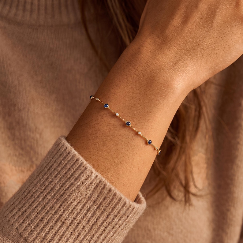 Puede incluir: Una delicada pulsera de cadena dorada con una serie de peque&ntilde;as piedras redondas de color azul oscuro y diminutas piedras blancas transparentes espaciadas uniformemente, engastadas en biseles dorados a lo largo de la cadena.