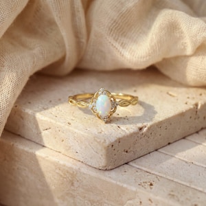 May include: A gold ring with an oval, white opal center stone. The opal is surrounded by small, clear gemstones and set in a decorative, filigree-style setting. The band features a delicate, leaf-like pattern.