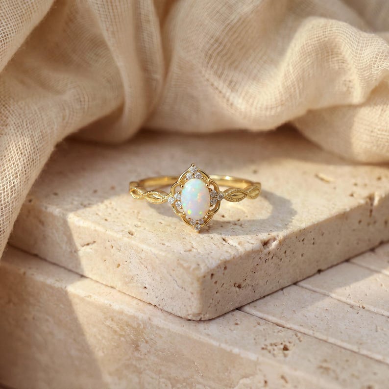 May include: A gold ring with an oval, white opal center stone. The opal is surrounded by small, clear gemstones and set in a decorative, filigree-style setting. The band features a delicate, leaf-like pattern.