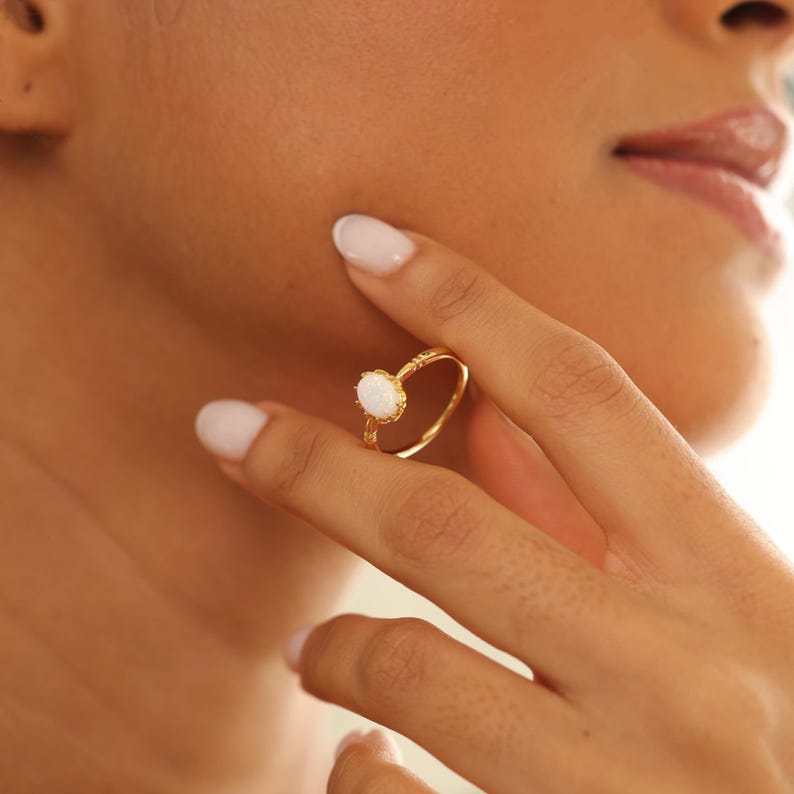 May include: A gold ring with a white gemstone. The ring is held up by a person's fingers. The ring has a delicate, ornate design around the band and the gemstone is oval-shaped.