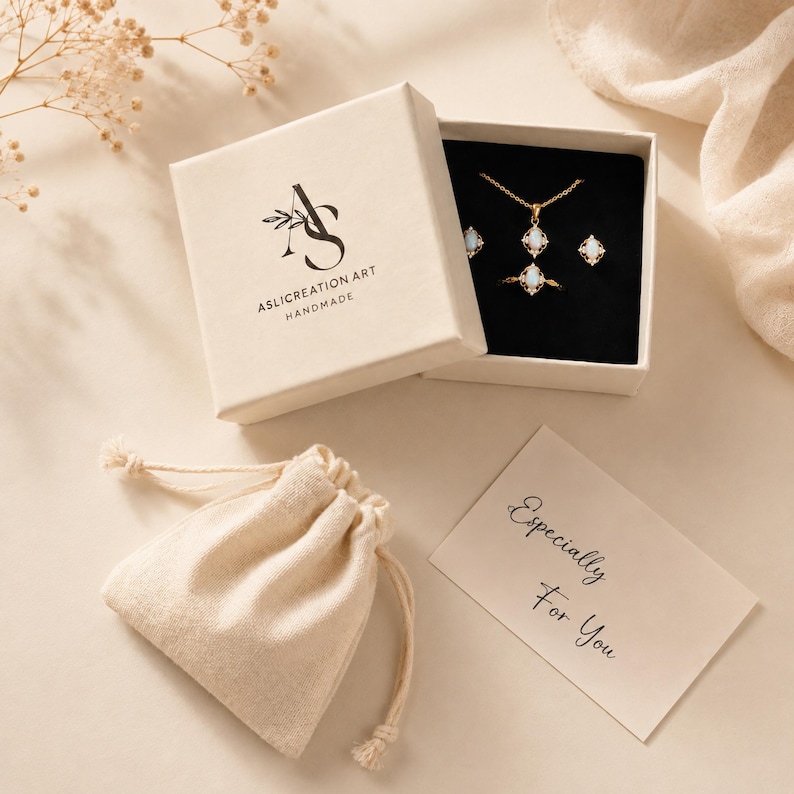 Puede incluir: Un conjunto de joyas con collar, anillo y pendientes dorados con piedras blancas ovaladas, en una caja beige con el texto ASLICREATION ART HANDMADE. Incluye una bolsa beige con cord&oacute;n y una tarjeta con el texto Especially For You.