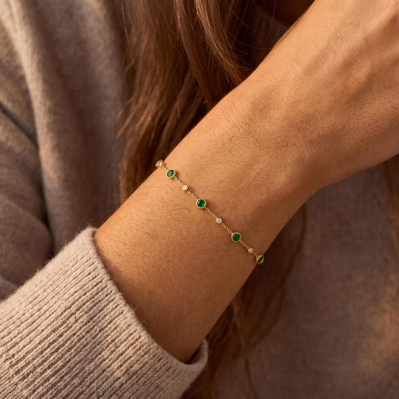 May include: A delicate gold chain bracelet featuring alternating round green gemstones and small clear white stones set in a bezel style. The bracelet is composed of a thin metallic link chain that wraps around the wrist, showcasing a repeating pattern.