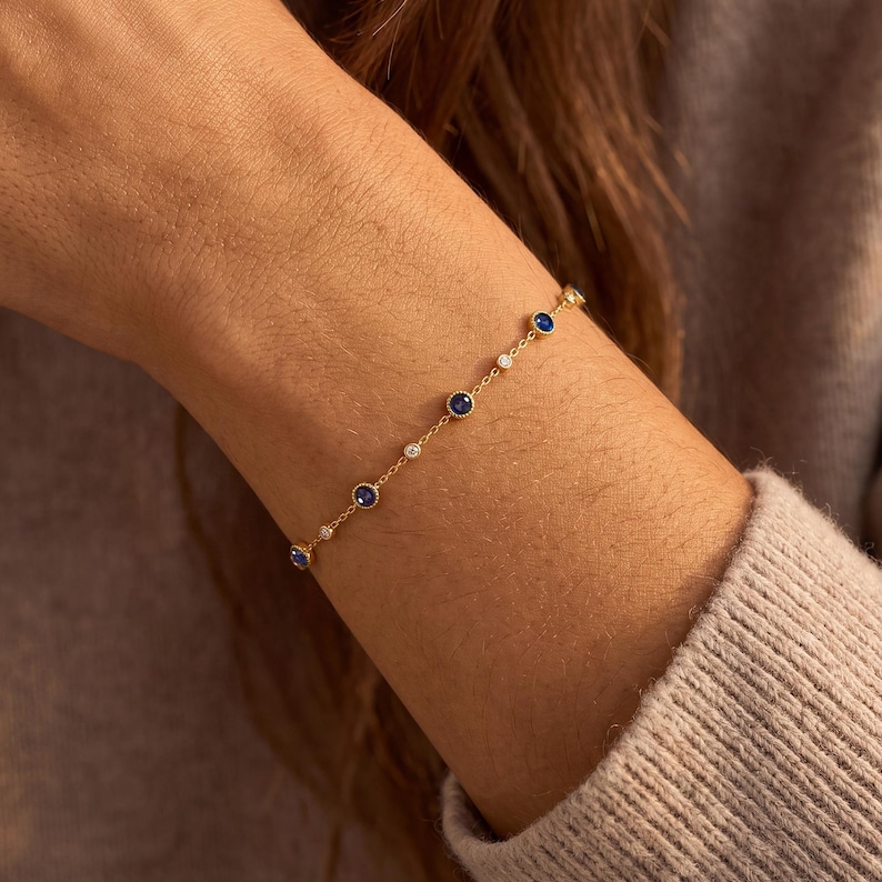 Puede incluir: Una delicada pulsera de cadena dorada que presenta piedras preciosas redondas de zafiro azul alternadas con piedras blancas transparentes m&aacute;s peque&ntilde;as, cada una engastada en un marco circular a lo largo de la cadena de eslabones.