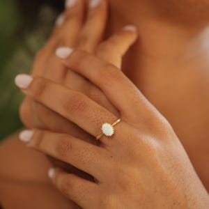 May include: A gold ring with an oval, white gemstone. The ring has a delicate, textured band and a decorative setting around the gemstone. The ring is on a person's finger.