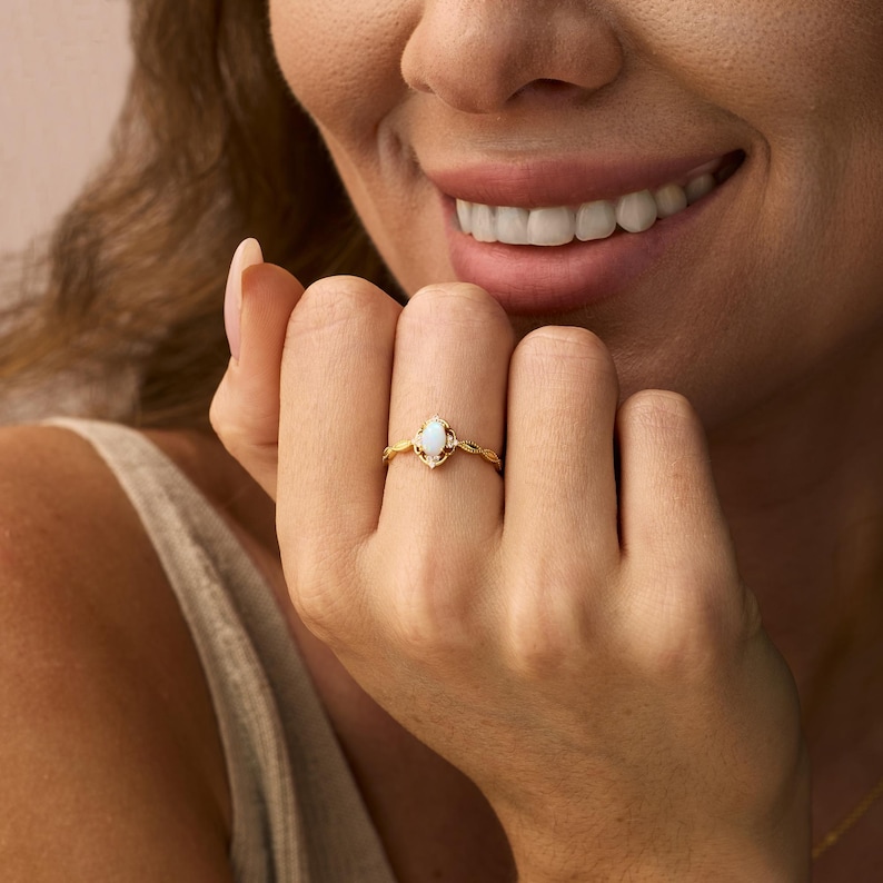 May include: A gold ring featuring an oval opal gemstone with small accent stones. The ring has a delicate, ornate design. The ring is on a person's finger.
