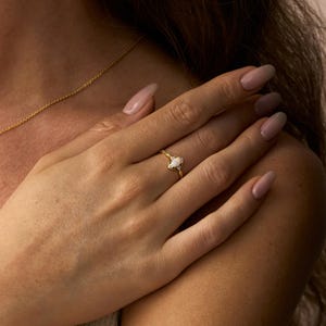 May include: A gold ring with a white oval-shaped gemstone. The ring is on a person's finger. The ring has a delicate, ornate design around the gemstone. A gold necklace is visible in the background.