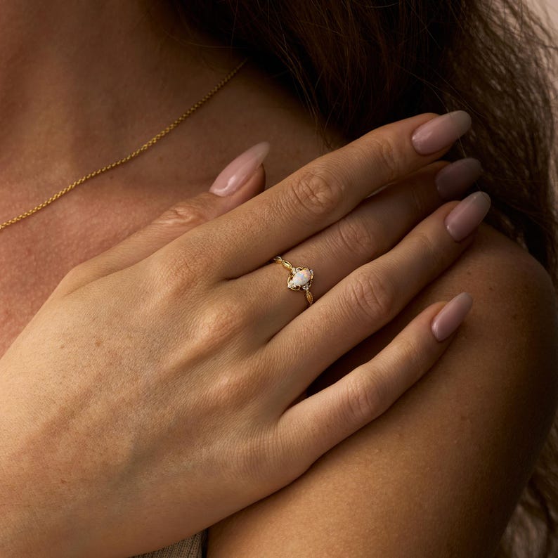 May include: A gold ring with a white oval-shaped gemstone. The ring is on a person's finger. The ring has a delicate, ornate design around the gemstone. A gold necklace is visible in the background.