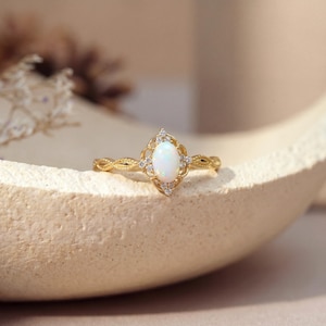 May include: A gold ring with an oval, white opal center stone. The ring features a decorative filigree design around the opal and along the band, with small accent stones. The ring is set against a neutral background.