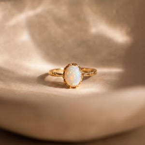 May include: A gold ring featuring an oval opal gemstone. The opal displays a milky white hue with iridescent flashes. The ring has an intricate setting and a slender band.
