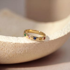 Puede incluir: Un anillo de oro con una serie de gemas de colores. El anillo presenta piedras verdes, rosas, azules, amarillas y rojas. El anillo está colocado sobre una superficie de tonos neutros.
