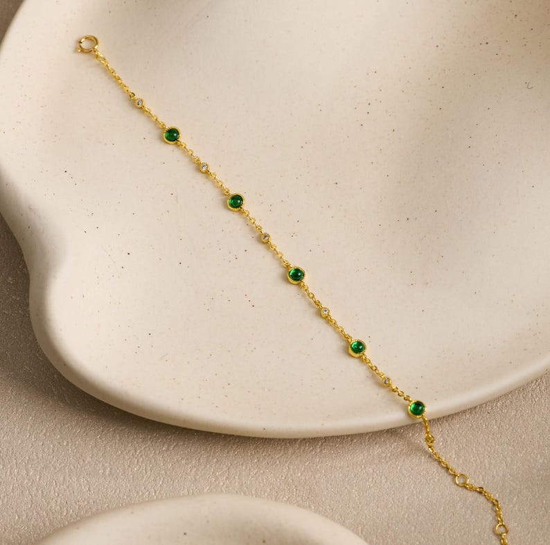 Puede incluir: Una delicada pulsera de cadena dorada que presenta una serie de piedras redondas de color verde vibrante espaciadas uniformemente, alternadas con piedras m&aacute;s peque&ntilde;as, claras y brillantes. La pulsera tiene una cadena fina y un cierre.