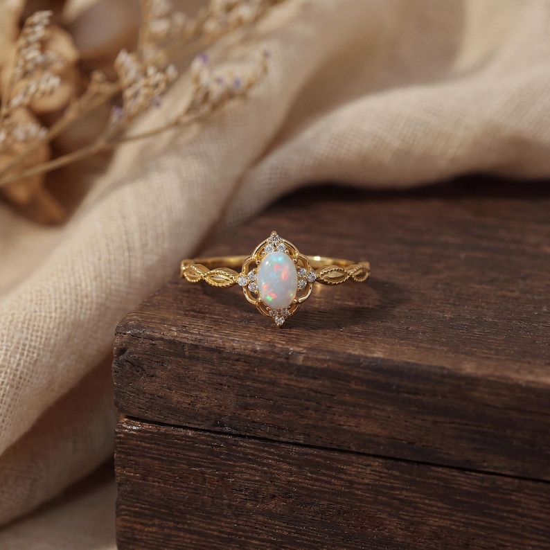 May include: A gold ring featuring an oval opal gemstone, surrounded by small diamonds. The ring has a delicate, ornate design with a textured band. The ring is displayed on a dark wooden surface, with a soft, neutral-colored fabric in the background.