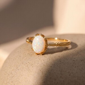 May include: A gold ring featuring an oval-shaped opal gemstone. The opal has a milky white color with iridescent flashes. The ring's band and setting are gold-colored, with a decorative design around the opal.