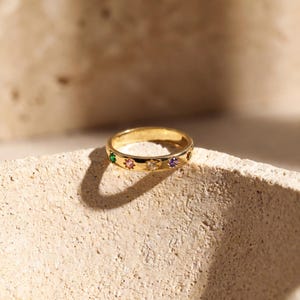 Könnte beinhalten: Ein goldener Ring mit einer Reihe farbiger Edelsteine. Der Ring weist ein sich wiederholendes Muster aus grünen, rosa, klaren und lila Steinen auf. Der Ring liegt auf einer strukturierten, hellfarbigen Oberfläche.