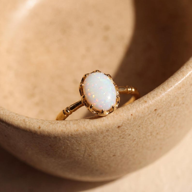 May include: A gold ring with an oval, white opal gemstone. The ring has a detailed, ornate setting and band. The opal has subtle pink and orange flecks. The ring is set against a neutral background, highlighting its elegant design.