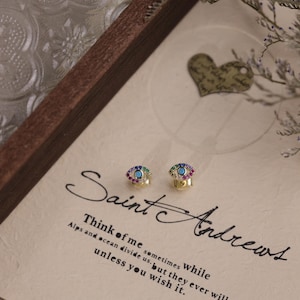 Puede incluir: Dos pendientes dorados con diseño de ojo, cada uno con un centro turquesa y detalles de piedras preciosas de colores. Los pendientes se muestran sobre un fondo con el texto "Saint Andrews" y una cita.
