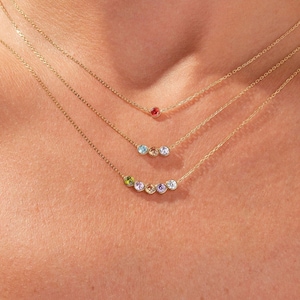 May include: Three delicate gold chain necklaces layered together. The top necklace features a single small red stone. The middle necklace has three small stones in blue, yellow, and blue. The bottom necklace has five stones in green, blue, yellow, blue, and white.