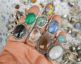 May include: A collection of silver rings with various gemstone inlays, including green malachite, blue labradorite, brown agate, and white moonstone. The rings are displayed on a hand.