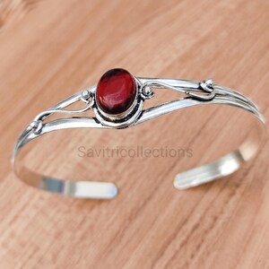 May include: A silver cuff bracelet with a central, oval, deep red gemstone. The open design features decorative swirls. The text "Savitricollections" is visible on the bracelet.