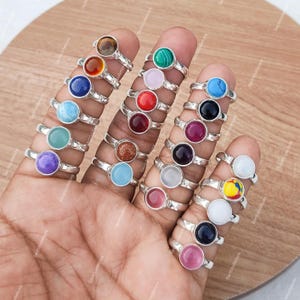 May include: A selection of silver rings, each with a different coloured gemstone. Gemstones include blue, green, red, purple, and other colours. The rings are displayed on fingers against a wooden surface.