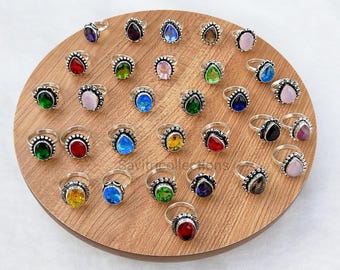May include: A collection of silver rings featuring teardrop-shaped gemstones in various colors including red, blue, green, yellow, and pink. Each ring has a beaded metal band design. The rings are arranged on a circular wooden display board.