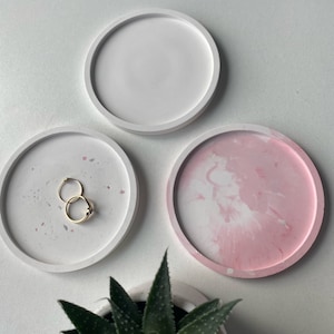 Könnte beinhalten: Drei runde Beton-Schmuckschalen. Eine Schale ist weiß, eine ist weiß mit rosa Flecken und eine ist rosa mit weißen Wirbeln. Zwei goldene Ringe liegen auf der weißen Schale mit rosa Flecken.