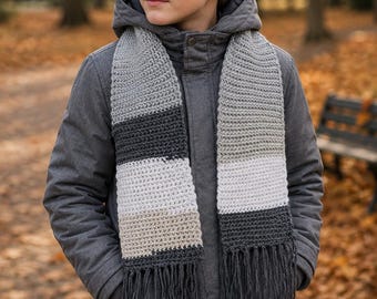 Könnte beinhalten: Ein gehäkelter Schal mit horizontalen Streifen in Anthrazit, Hellgrau, Weiß und Beige, der an beiden Enden mit langen anthrazitfarbenen Fransen versehen ist und über einer anthrazitfarbenen Kapuzenjacke mit Reißverschluss getragen wird.