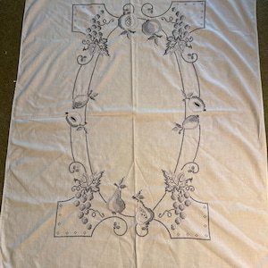 May include: A rectangular white fabric tablecloth featuring a central cross-stitch embroidery design in dark gray thread. The pattern consists of a decorative border of grapes, leaves, and various fruits arranged in an oval shape on the fabric.