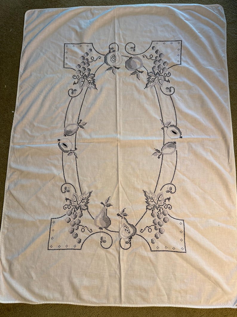 May include: A rectangular white fabric tablecloth featuring a central cross-stitch embroidery design in dark gray thread. The pattern consists of a decorative border of grapes, leaves, and various fruits arranged in an oval shape on the fabric.
