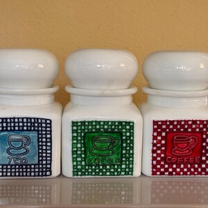 May include: Three white ceramic canisters with matching lids. Each canister has a square label with a different color and the name of a beverage: tea, cocoa, and coffee. The tea label is blue, cocoa is green, and coffee is red.