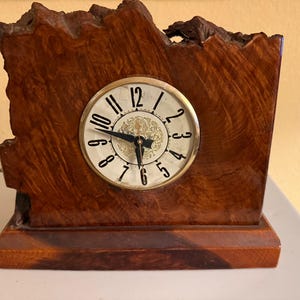 May include: A wooden mantel clock with a round clock face. The clock face has black numerals and hands against a cream background. The clock is set in a dark brown, textured wooden frame.