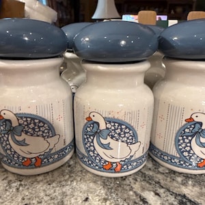 May include: A set of white ceramic canisters with blue lids, each featuring a graphic of a white goose with an orange beak and feet, wearing a blue bow. The canisters have a blue floral circular pattern and vertical blue pinstripes on the body.