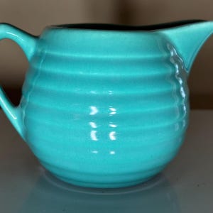 May include: A turquoise ceramic creamer pitcher featuring a rounded, bulbous body with horizontal ribbed detailing, a curved handle on the left side, and a pointed pouring spout on the right side, finished with a smooth, glossy glaze.