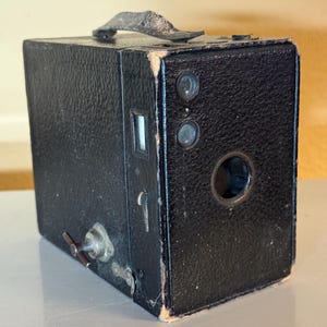 May include: A vintage black box camera with a leather handle. The camera has a rectangular shape with a lens, viewfinder, and various dials and levers. The camera is likely from the early to mid-20th century.
