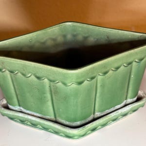 May include: A green ceramic planter with a matching saucer. The planter has a diamond shape with scalloped edges and vertical ribbed sides. The saucer is also diamond-shaped with a scalloped edge. A decorative item for plants.