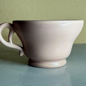 May include: A pale pink ceramic teacup with a curved handle and a flared rim. The cup has a rounded base and a smooth, matte finish. The handle is a unique, open design.