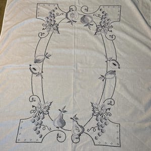 May include: A white rectangular tablecloth featuring a central cross-stitch embroidery pattern in dark gray thread. The design consists of a decorative border of grapes, pears, and pomegranates arranged in an oval shape with scrolling vines.