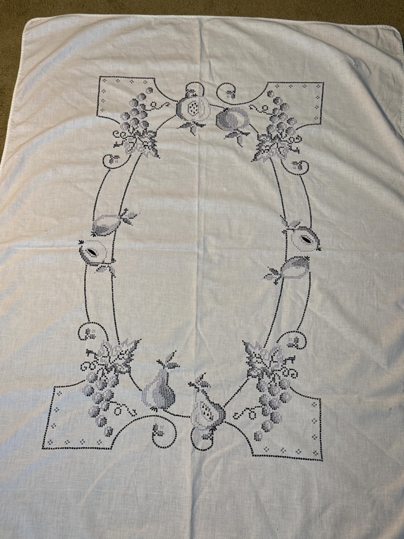 May include: A white rectangular tablecloth featuring a central cross-stitch embroidery pattern in dark gray thread. The design consists of a decorative border of grapes, pears, and pomegranates arranged in an oval shape with scrolling vines.