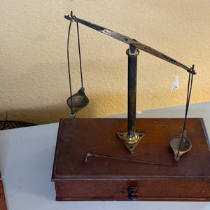 May include: An antique balance scale with a dark wooden base and a brass and metal arm. The scale has two hanging pans and a small drawer. A metal tool rests on the base.