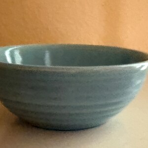 May include: A light blue ceramic bowl with a smooth, glossy interior and a textured exterior featuring subtle horizontal ridges. The bowl has a rounded shape with a slightly flared rim and a consistent, muted blue color throughout the entire piece.