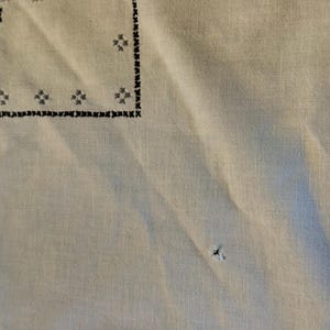 May include: A light beige fabric featuring black cross-stitch embroidery in a geometric border pattern. The textile displays a visible small hole in the lower section and a faint stain near the upper right corner, with a textured weave throughout.