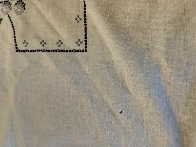 May include: A light beige fabric featuring black cross-stitch embroidery in a geometric border pattern. The textile displays a visible small hole in the lower section and a faint stain near the upper right corner, with a textured weave throughout.