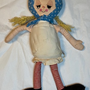 May include: A vintage handmade doll with a painted face, yellow yarn pigtails, and a blue patterned headscarf. The doll wears a cream-colored dress, red and white striped stockings, and black shoes. The doll's arms are outstretched.