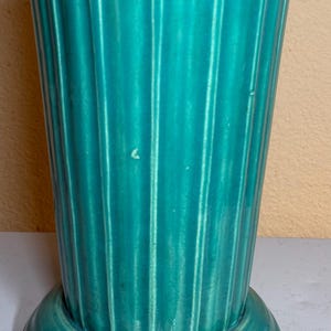 May include: A teal-colored ceramic vase with vertical ribbed detailing. The vase has a flared top and a circular base. The glossy finish reflects light, highlighting the texture. A decorative piece for home decor.