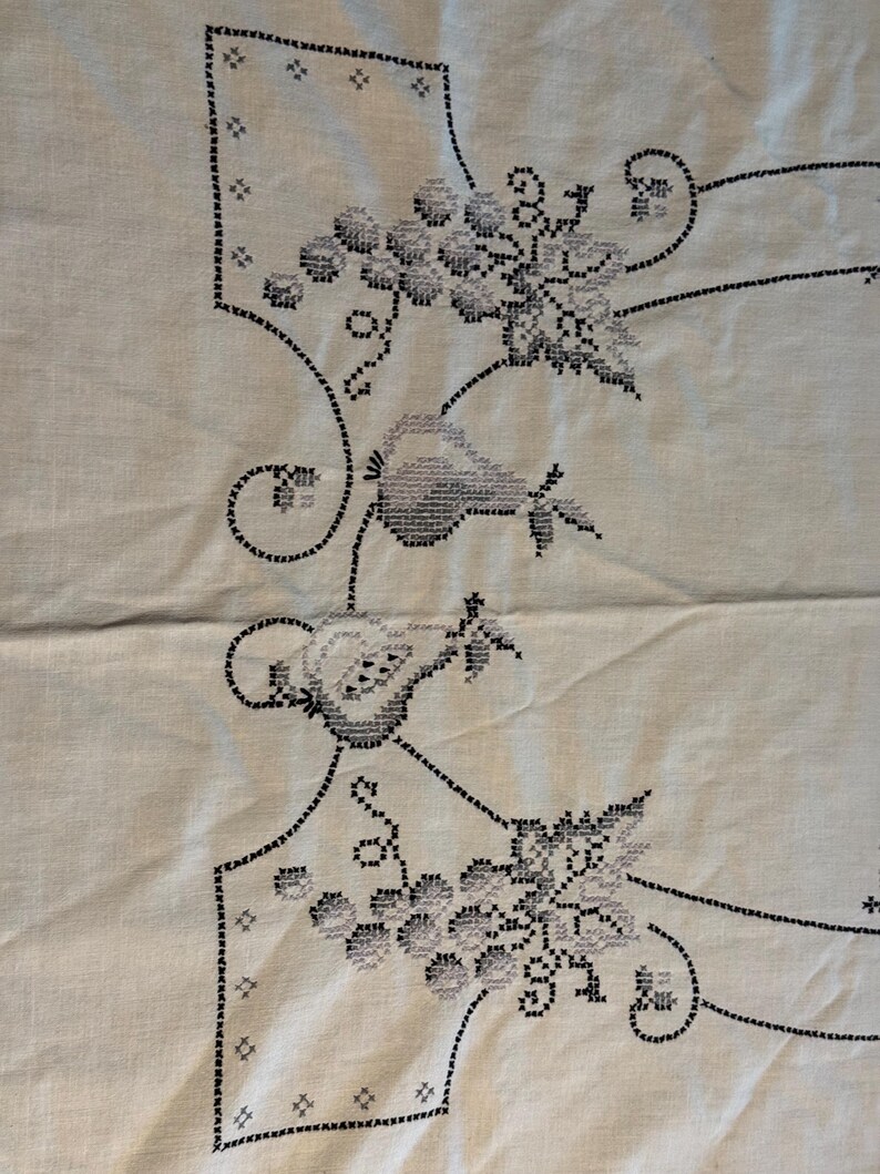 May include: A light-colored fabric featuring black cross-stitch embroidery. The design displays clusters of grapes and pear-shaped fruits connected by thin, curved lines. The embroidery is arranged in a symmetrical pattern with decorative borders.